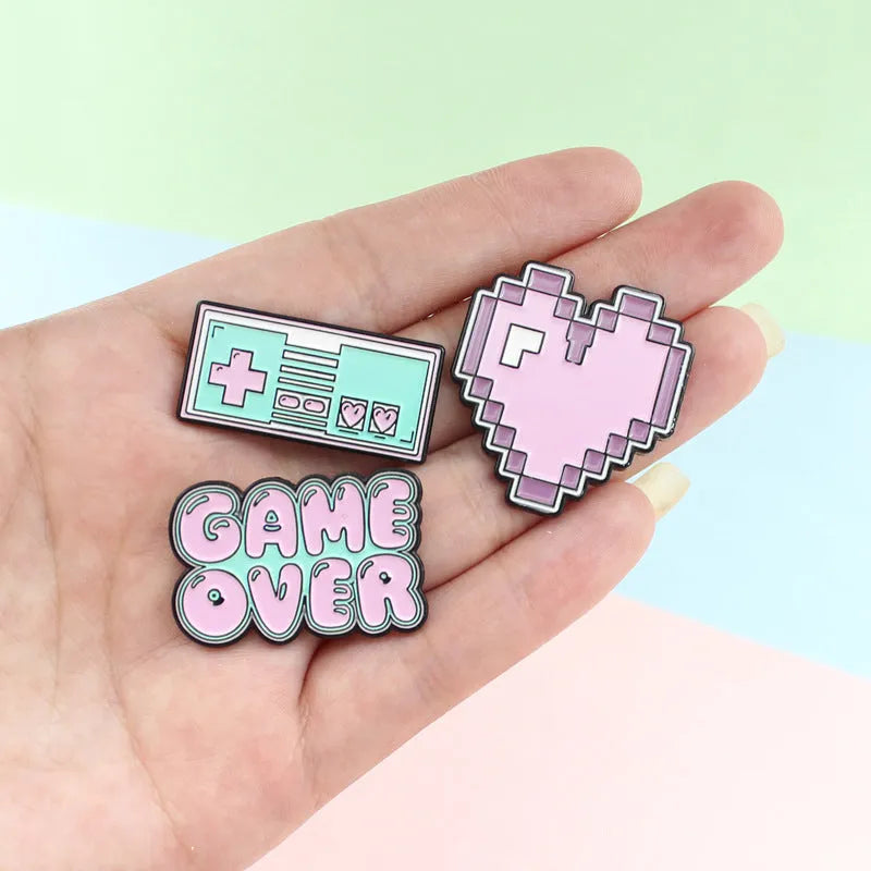 A metal alloy game console badge shown in one of three available colours, suitable for attaching to bags.