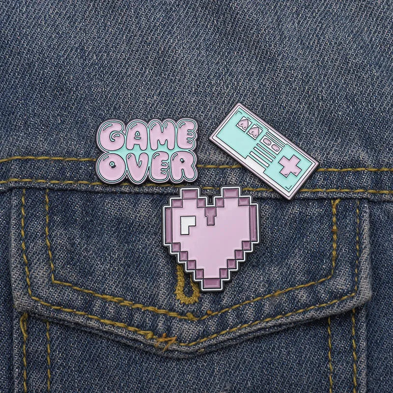 A metal alloy game console badge shown in one of three available colours, suitable for attaching to bags.