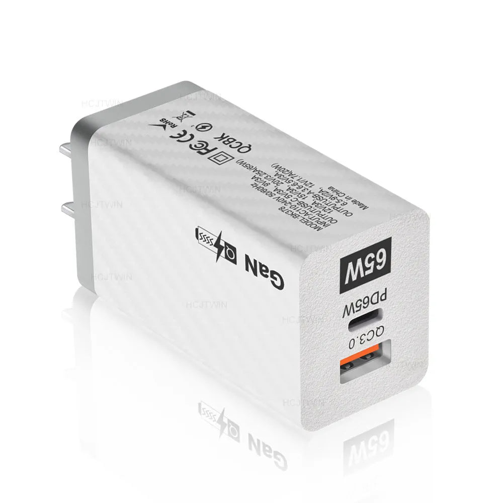 Compact white 65W Gallium Nitride laptop charger with UK, US and EU plug attachments on a desk.
