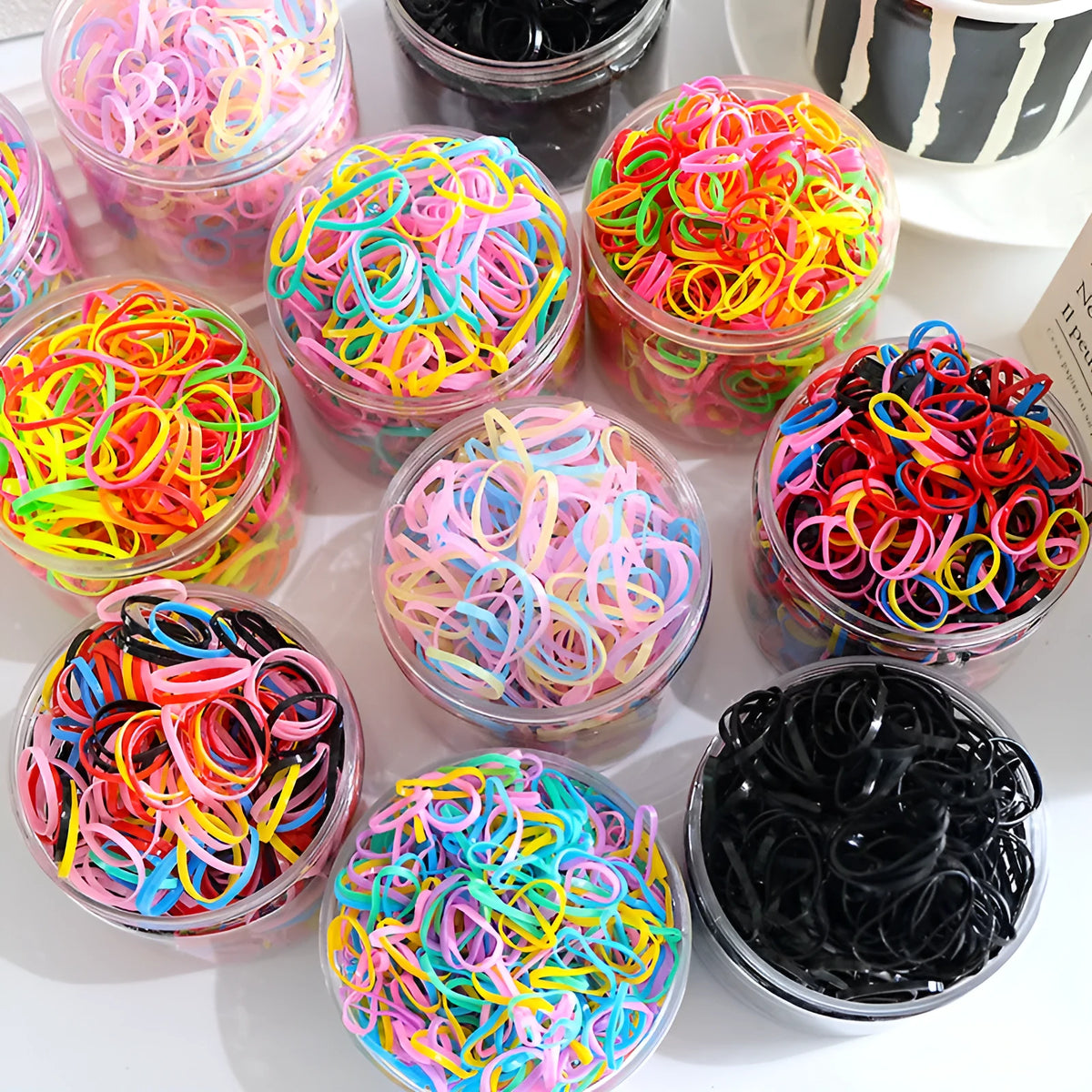 Assortment of colourful small TPU baby hair ties in a reusable plastic container.