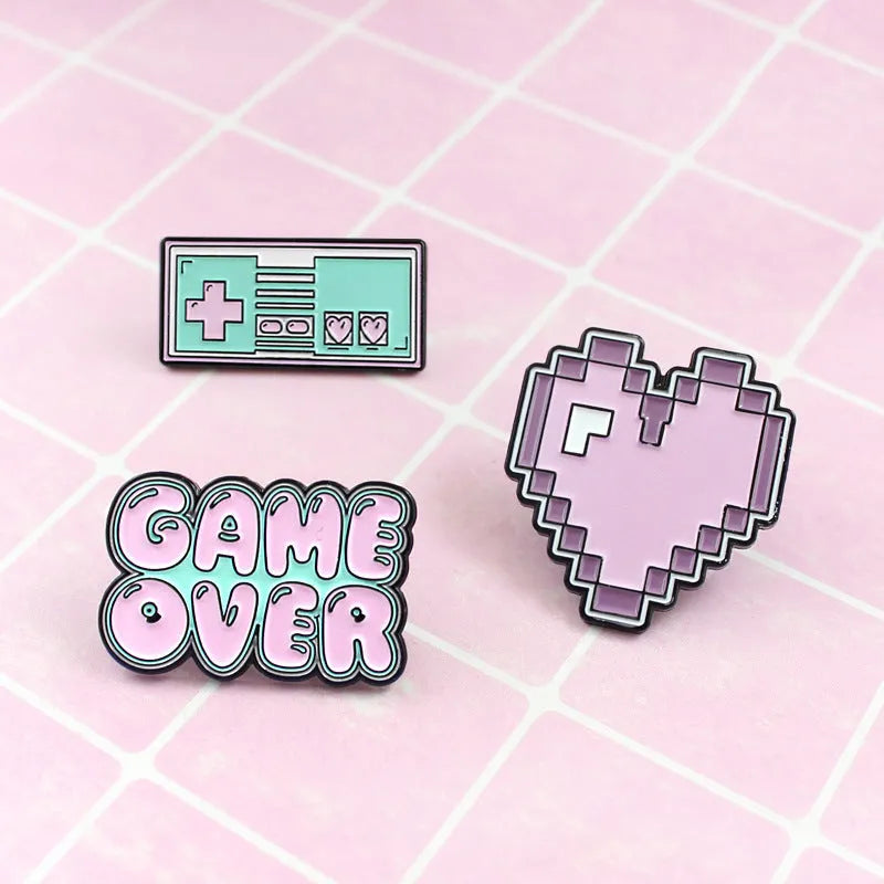 A metal alloy game console badge shown in one of three available colours, suitable for attaching to bags.