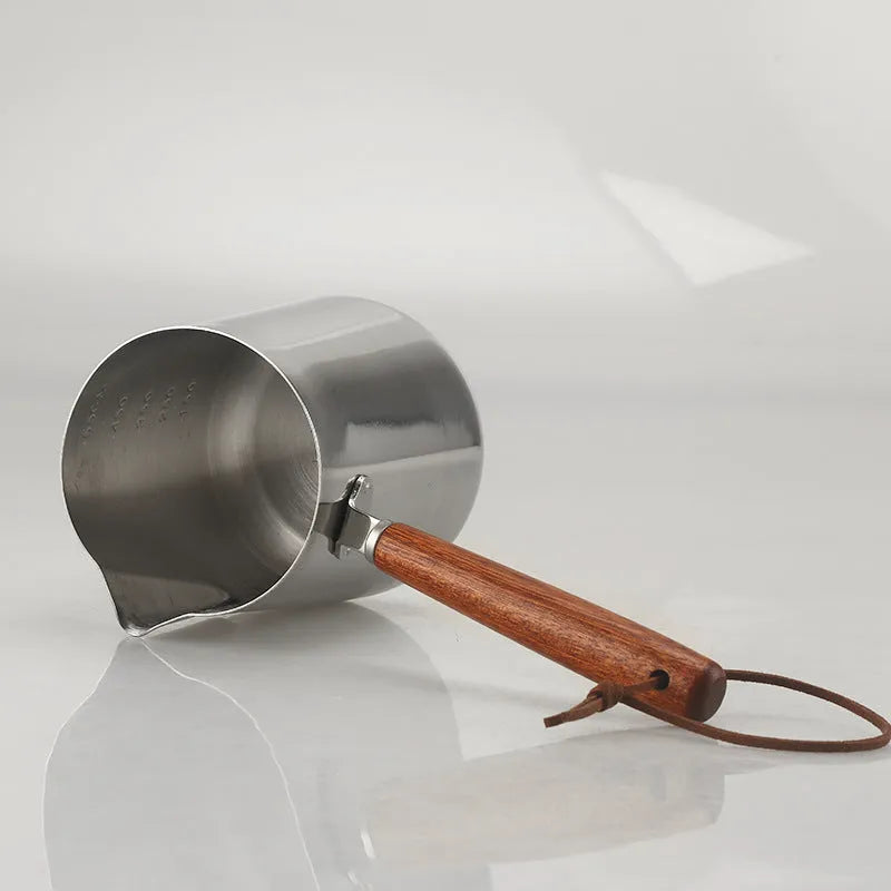 Stainless steel milk jug with measurement scale and wooden handle on a counter.
