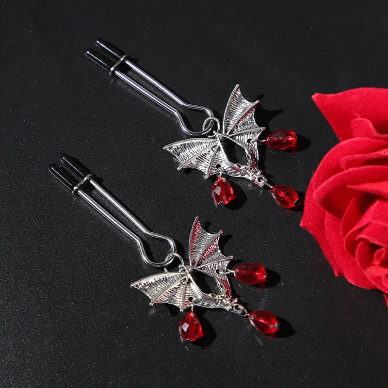 A pair of silver metal bat-shaped adjustable nipple clamps on a plain background.