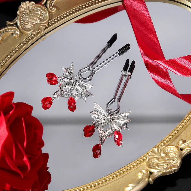 A pair of silver metal bat-shaped adjustable nipple clamps on a plain background.