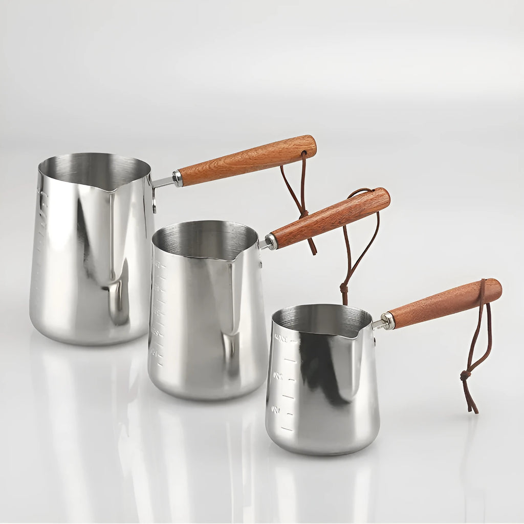 Stainless steel milk jug with measurement scale and wooden handle on a counter.