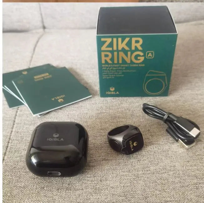 Zikr1 smart ring in silver on a charging cable next to its case, showing the OLED display.