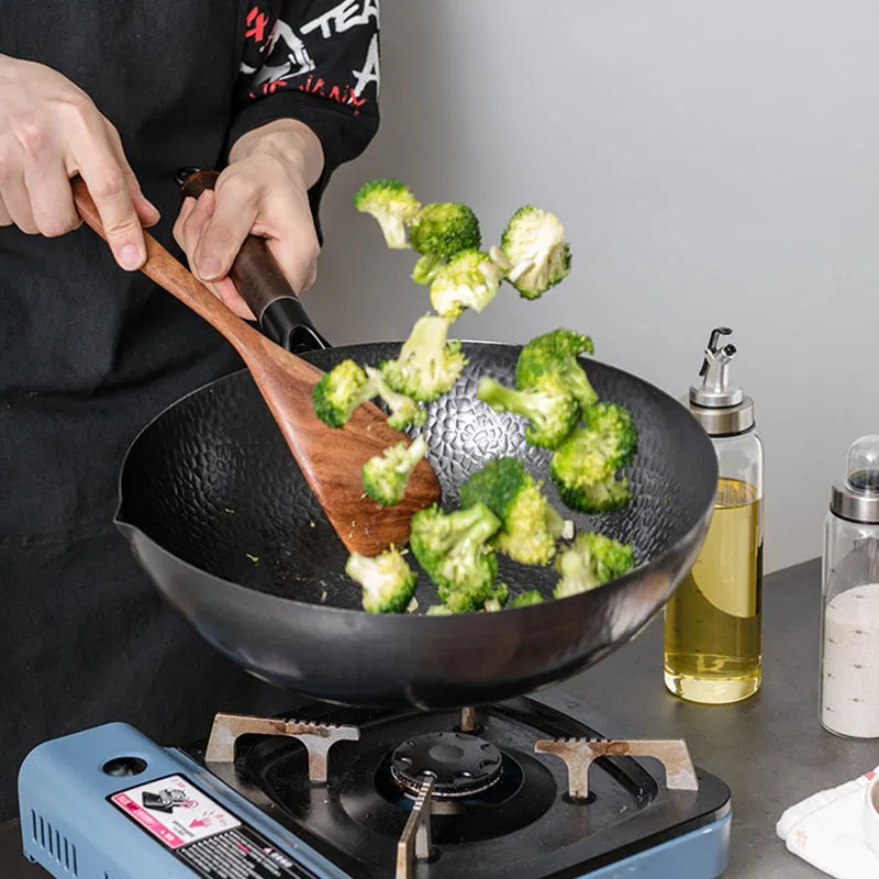 Traditional black iron wok pan with a wooden handle on a hob.