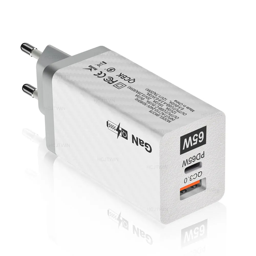 Compact white 65W Gallium Nitride laptop charger with UK, US and EU plug attachments on a desk.